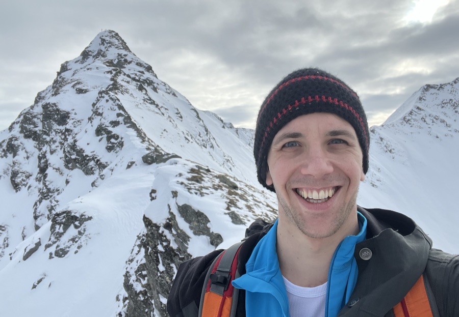 Dominik on a snowy mountain ridge, smiling, with a peak behind him.